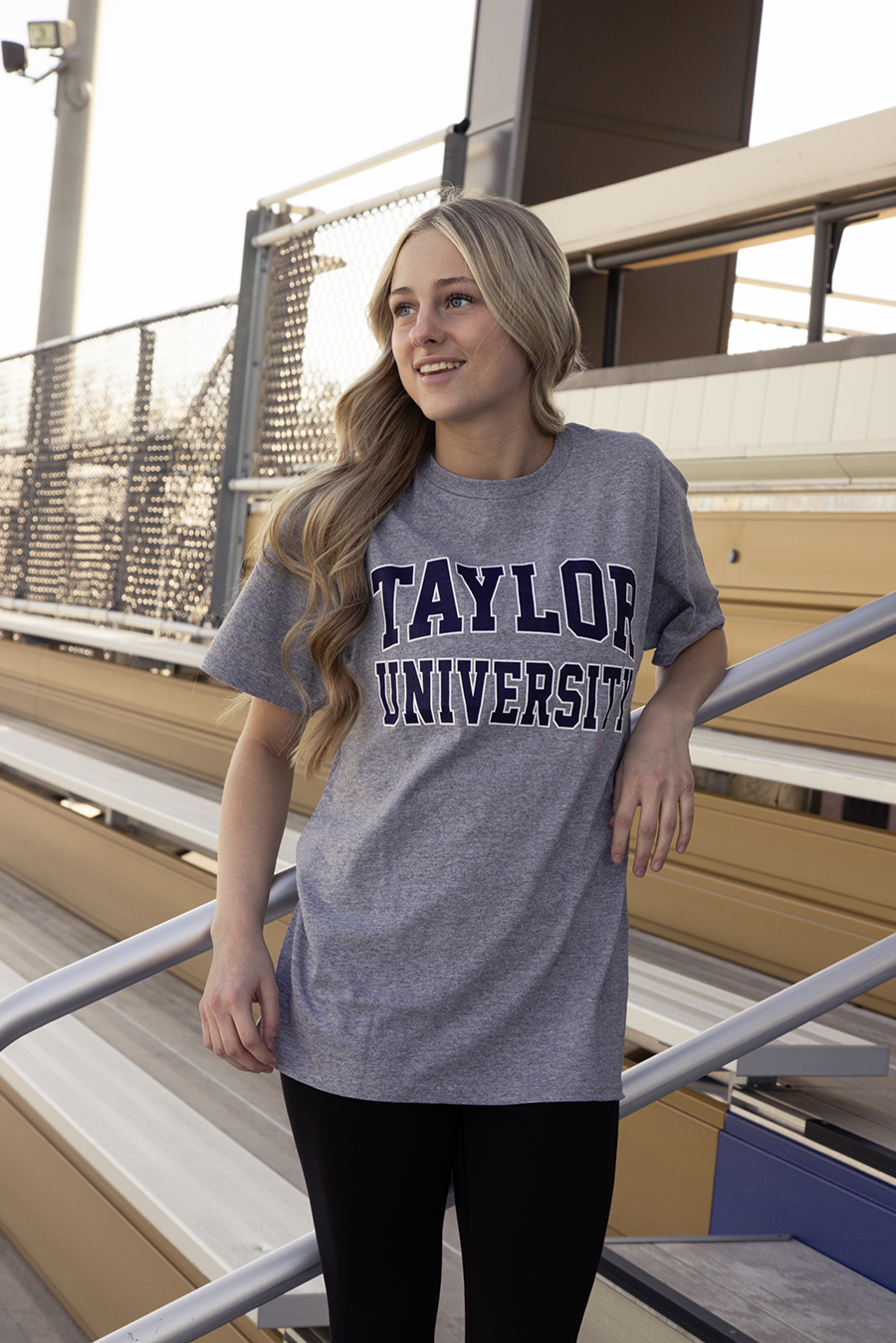 Girl standing on the bleachers wearing a gray short sleeve Taylor University shirt.