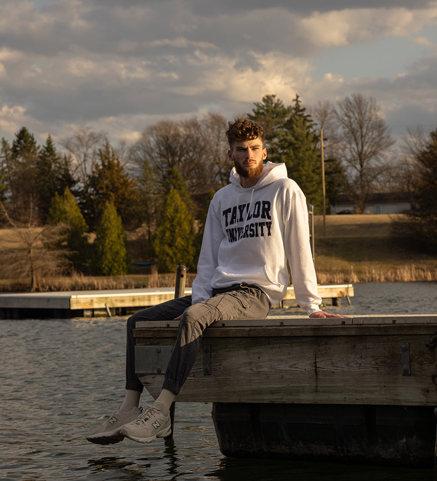 Male sitting on a dock in the water wearing a Taylor University hoodie.