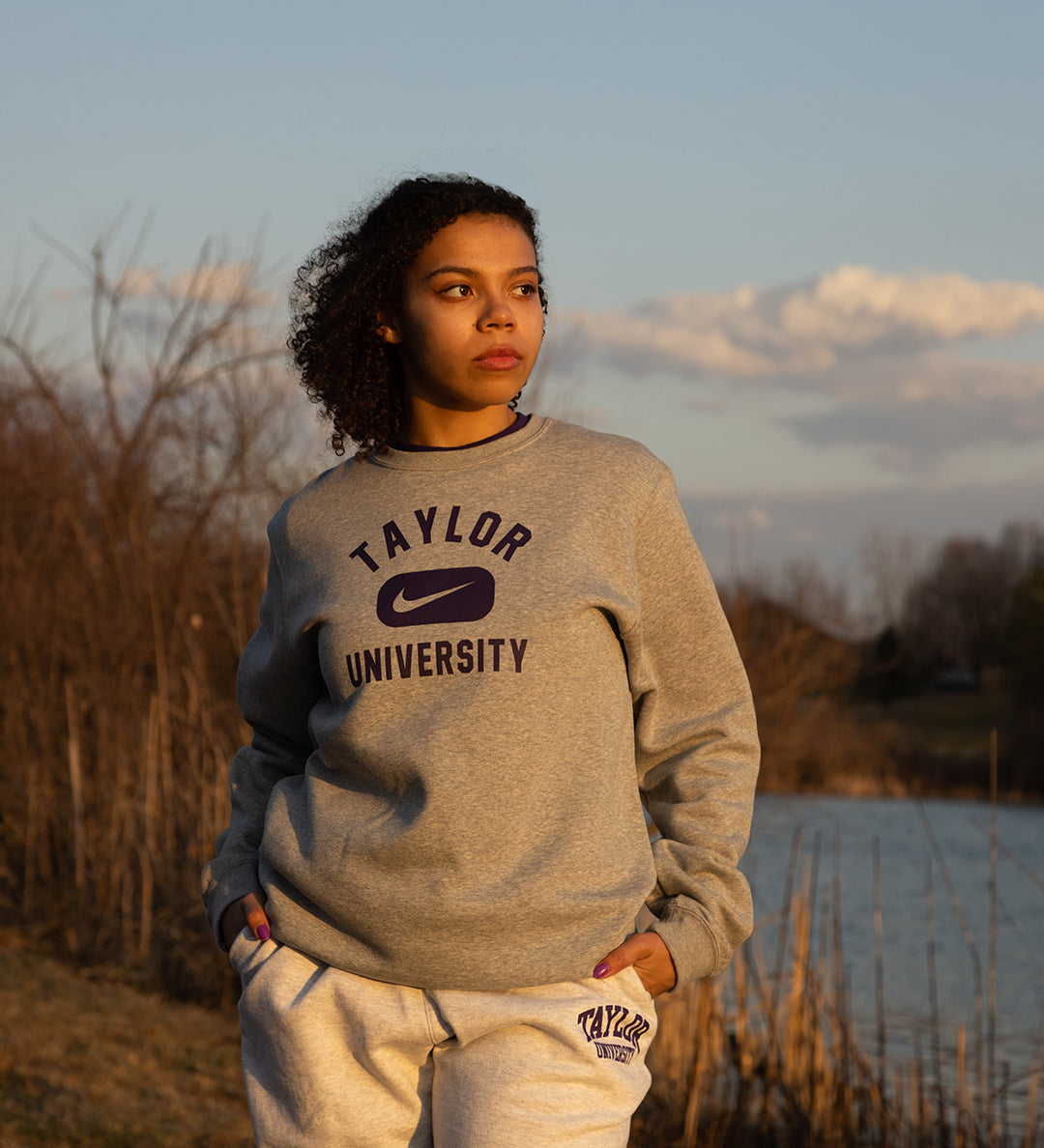 Girl posing in Taylor University apparel with trees and pond behind her.