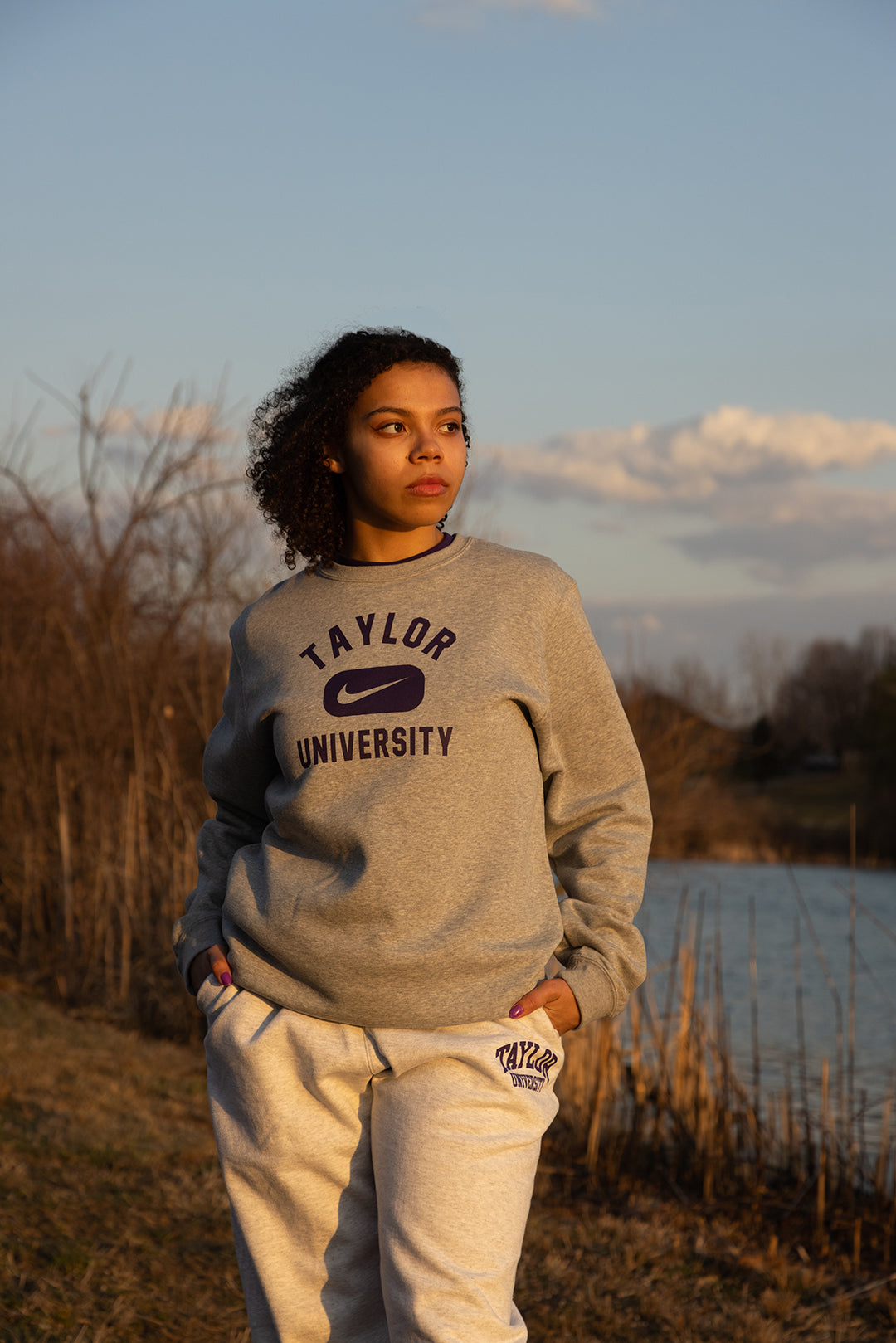 Girl posing in Taylor University apparel with trees and pond behind her.