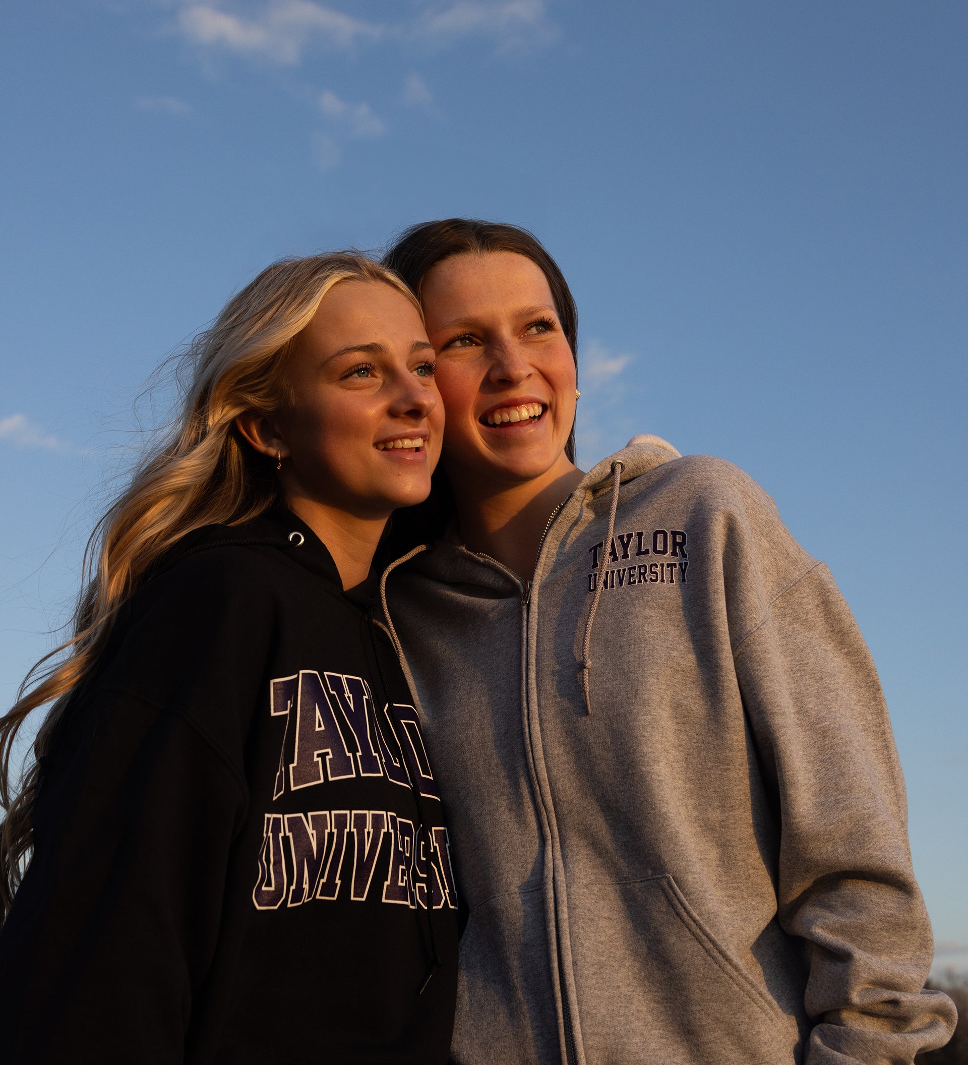 2 Girls posing with the sky behind them wearing Taylor University hoodies. 