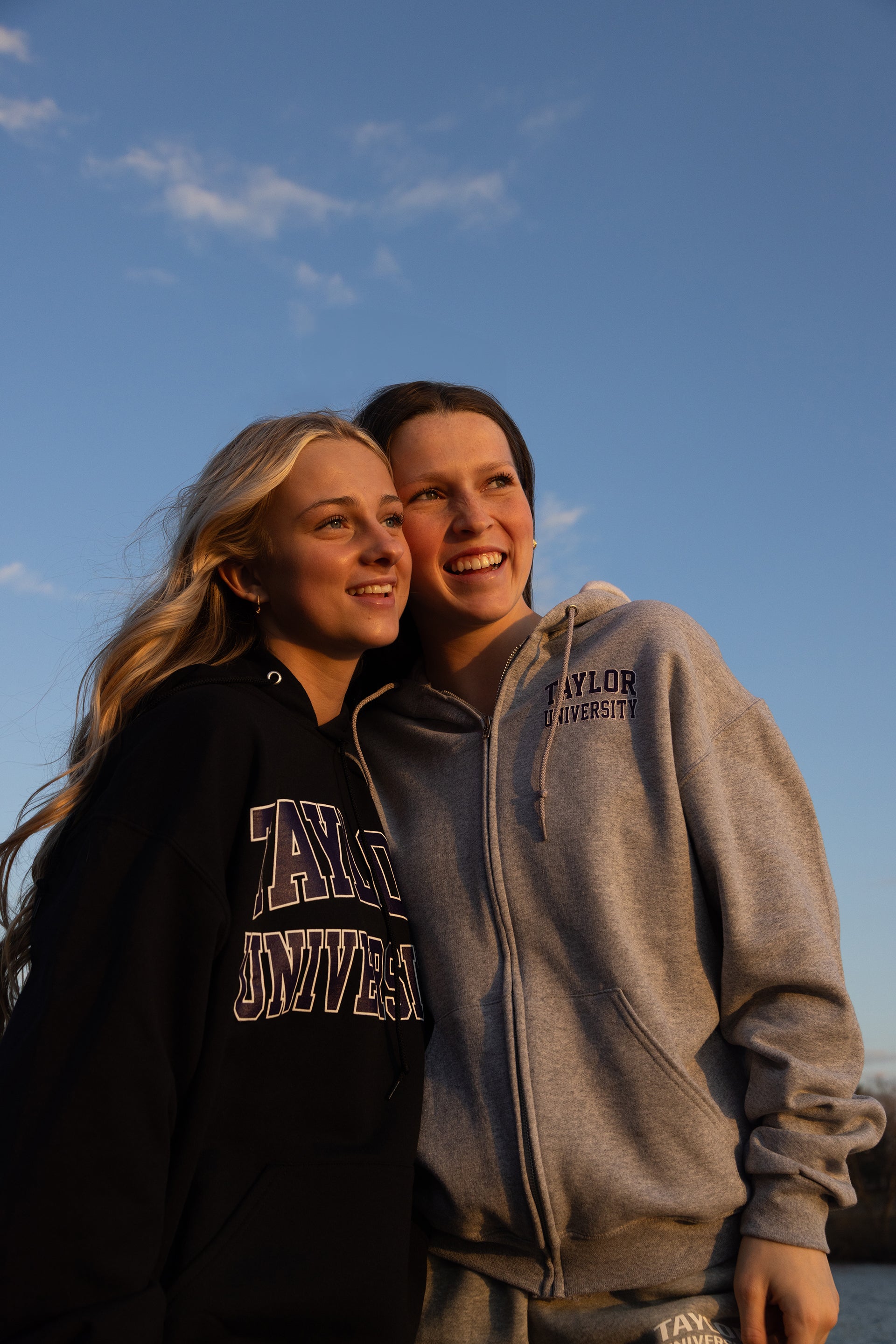 2 Girls posing with the sky behind them wearing Taylor University hoodies. 